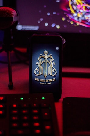 A smartphone displays an ornate logo with a crown at the top and ornate lettering below reading 'The King of Blutz'. The phone is set in a dimly lit environment with a microphone to the left and a keyboard in the foreground, featuring colored lights. The background suggests a computer monitor with colorful abstract graphics.