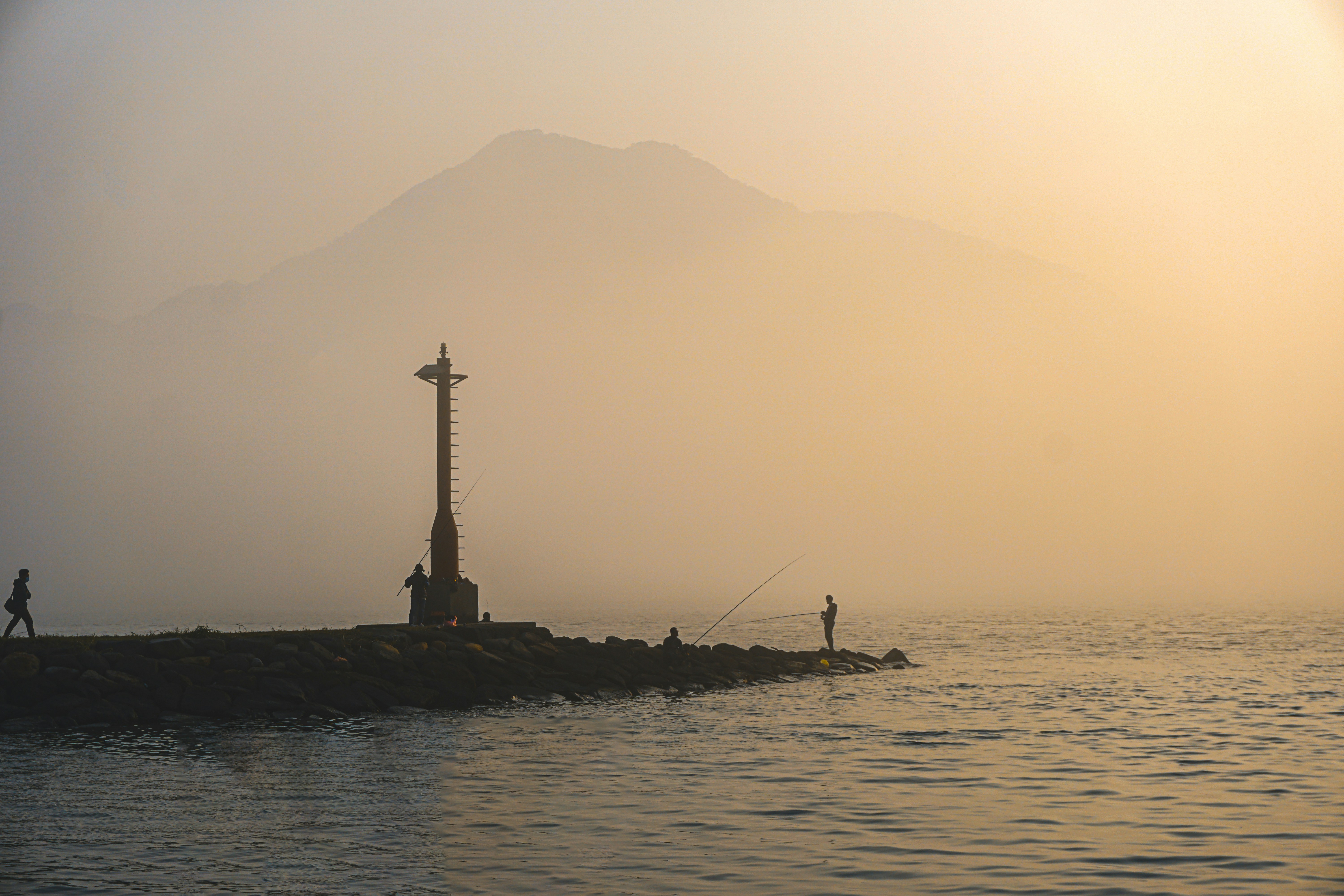 Silhouettes of a beacon and anglers on a rocky jetty glow in warm sunrise light over calm water, with hazy mountain silhouettes in the background.