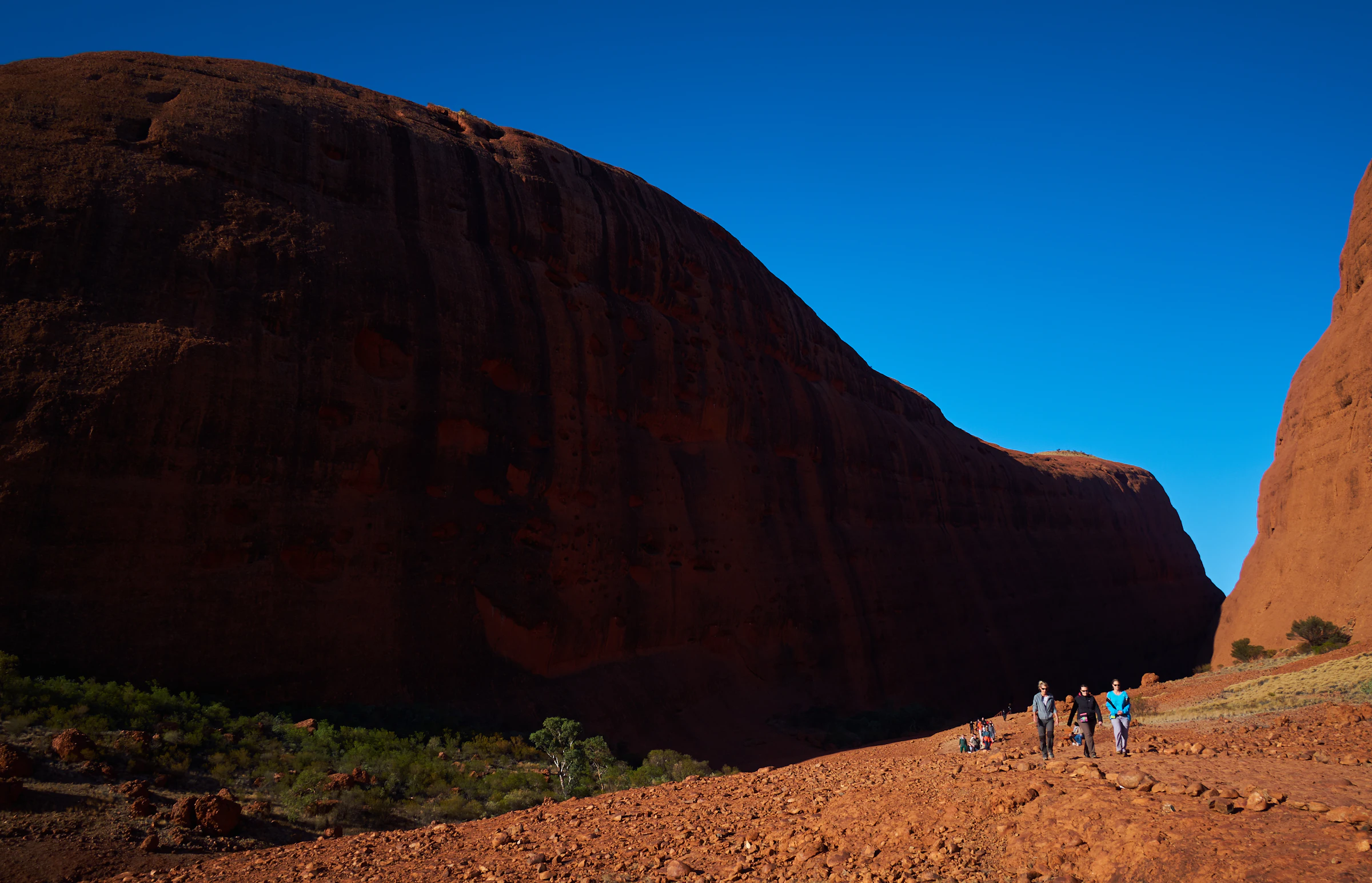 Close view of towering dark-red rock walls with visitors walking along a rocky trail at the base.