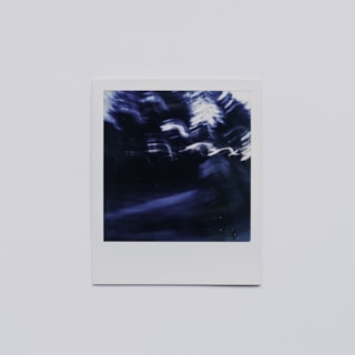 A blurry, abstract photograph with dark, indistinct shapes and white and blue color streaks. The image is framed within a white border, giving it a classic Polaroid look.