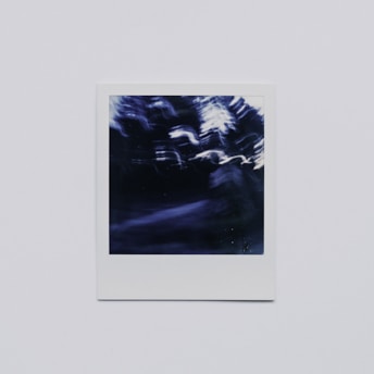 A blurry, abstract photograph with dark, indistinct shapes and white and blue color streaks. The image is framed within a white border, giving it a classic Polaroid look.