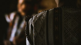 A close-up view of a textured, patterned fabric, possibly part of a traditional garment, with muted lighting creating a warm and intimate atmosphere. The design features geometric ornamental patterns with fine details.