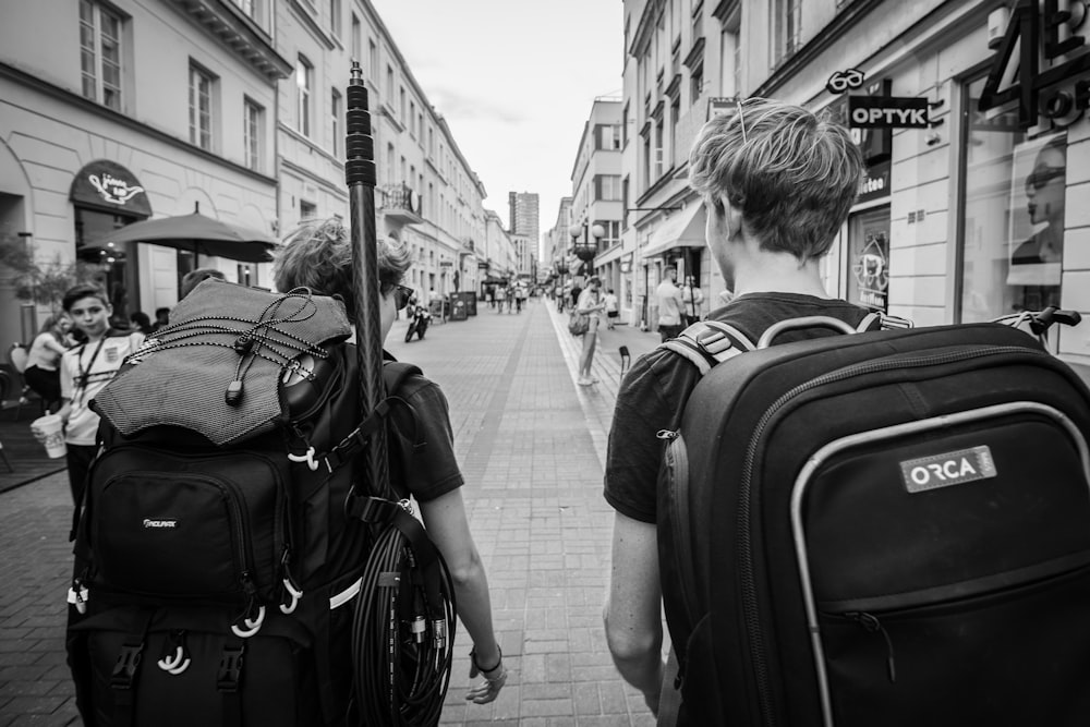 grayscale photo of man in black jacket carrying backpack walking on sidewalk