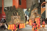 Two performers wearing traditional costumes featuring large, ornate lion masks adorned with peacock feathers. The costumes have vibrant yellow, red, and orange fabric accents. In the background, spectators, including children and adults, watch the performance.