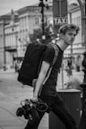A black and white image of a person walking down a street carrying a large backpack and camera equipment. The person has a pensive expression and is wearing casual clothing. In the background, there are buildings lining the street, and a taxi sign is visible.