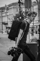 A black and white image of a person walking down a street carrying a large backpack and camera equipment. The person has a pensive expression and is wearing casual clothing. In the background, there are buildings lining the street, and a taxi sign is visible.