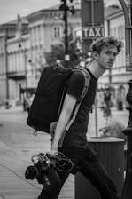 A black and white image of a person walking down a street carrying a large backpack and camera equipment. The person has a pensive expression and is wearing casual clothing. In the background, there are buildings lining the street, and a taxi sign is visible.