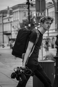 A black and white image of a person walking down a street carrying a large backpack and camera equipment. The person has a pensive expression and is wearing casual clothing. In the background, there are buildings lining the street, and a taxi sign is visible.