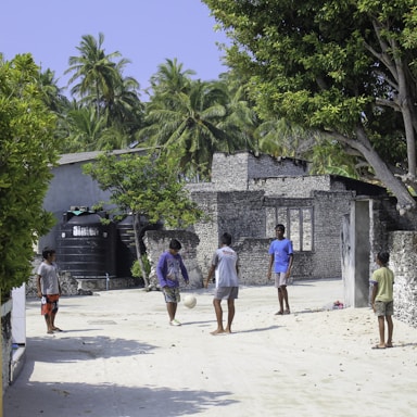 Several children are playing soccer on a sandy street, surrounded by lush green trees and rustic buildings made of stone bricks. The environment is tropical, with palm trees visible in the background under a clear blue sky. The children are casually dressed and the setting appears to be a quiet, rural area.