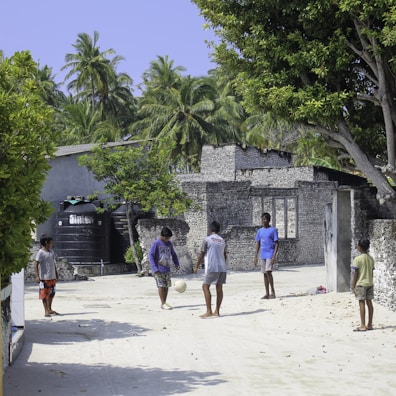 Several children are playing soccer on a sandy street, surrounded by lush green trees and rustic buildings made of stone bricks. The environment is tropical, with palm trees visible in the background under a clear blue sky. The children are casually dressed and the setting appears to be a quiet, rural area.
