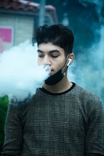 A person with dark hair is exhaling a large cloud of vapor while wearing a face mask pulled below the chin and earbuds. The individual is dressed in a plaid-patterned sweater and is positioned against an outdoor background obscured by vapor.