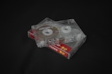 Two transparent cassette tapes are placed on a dark surface. The tapes are stacked on top of each other at an angle, revealing the spools and tape inside. The bottom cassette has a label with a red and yellow design stating 'Ferro 90'.