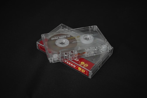 Two transparent cassette tapes are placed on a dark surface. The tapes are stacked on top of each other at an angle, revealing the spools and tape inside. The bottom cassette has a label with a red and yellow design stating 'Ferro 90'.