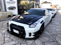 A modified sports car is showcased, featuring a sleek black and white exterior with custom detailing and branding. It is parked on a stone-paved area near other sports cars, with a building in the background adorned with automotive posters. A palm tree is visible beside the structure.