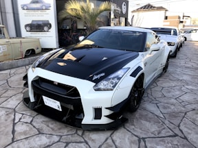 A modified sports car is showcased, featuring a sleek black and white exterior with custom detailing and branding. It is parked on a stone-paved area near other sports cars, with a building in the background adorned with automotive posters. A palm tree is visible beside the structure.
