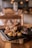 A cast iron pan filled with grilled meat and caramelized onions rests on a wooden platter, set on a rustic wooden table. The background features blurred lights and furniture, creating a cozy, ambient atmosphere.