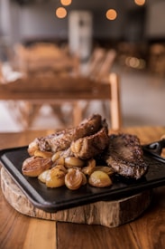 A cast iron pan filled with grilled meat and caramelized onions rests on a wooden platter, set on a rustic wooden table. The background features blurred lights and furniture, creating a cozy, ambient atmosphere.