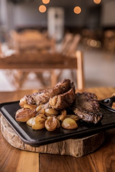 A cast iron pan filled with grilled meat and caramelized onions rests on a wooden platter, set on a rustic wooden table. The background features blurred lights and furniture, creating a cozy, ambient atmosphere.