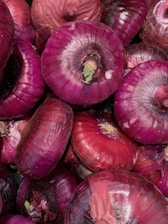 A close-up of multiple red onions with a purple hue, displaying their textured skins and green sprouts emerging from some. The onions are densely packed and occupy the entire frame.