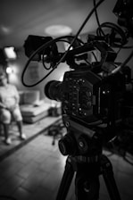 A black and white image showing a professional video camera on a tripod in the foreground, with its intricate buttons and wires visible. In the background, slightly out of focus, there is a person sitting on a sofa in a well-lit room, with what appears to be additional lighting equipment visible.