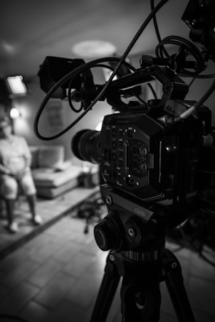 A black and white image showing a professional video camera on a tripod in the foreground, with its intricate buttons and wires visible. In the background, slightly out of focus, there is a person sitting on a sofa in a well-lit room, with what appears to be additional lighting equipment visible.