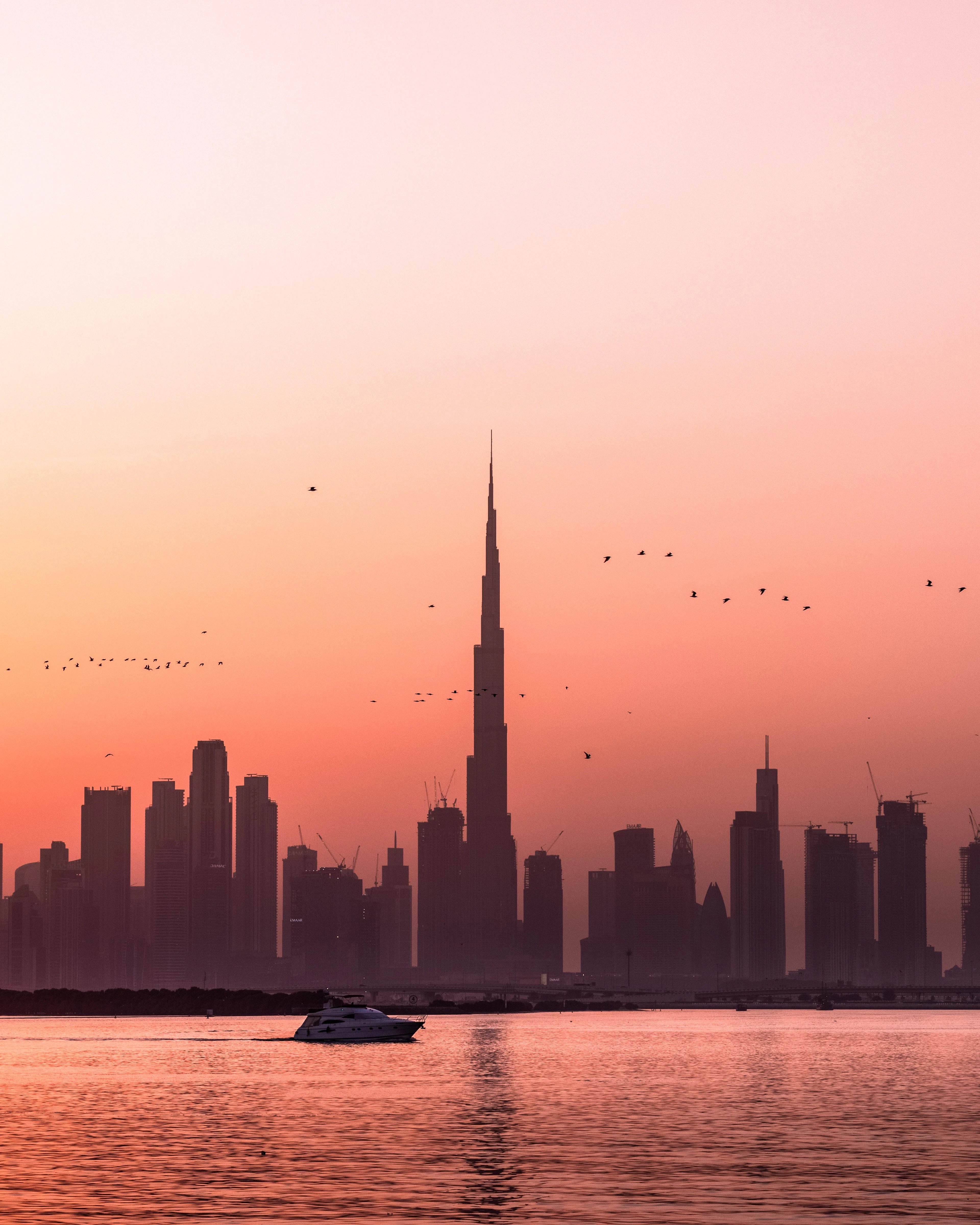 The Dubai skyline and Burj Khalifa stand in silhouette against a coral sunset with birds over the water.
