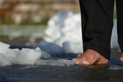 A person's bare foot is seen walking on a surface covered with patches of melting ice. The individual is wearing black pants, and the scene captures a close-up view of the foot interacting with the wet ground and crushed ice.