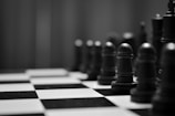 A monochrome image of a chessboard with a close-up focus on chess pieces. The pawns and other pieces are positioned on the checkered board, creating a sense of strategy and contemplation.