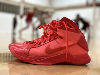 A pair of bright red basketball shoes is placed on a glossy indoor court. The background is blurred, highlighting several players engaged in a game, which adds depth to the scene.
