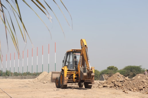 A construction site featuring a yellow backhoe loader parked amidst piles of dirt. In the background stand tall poles painted in the colors of the Indian flag: orange, white, and green. The scene is set on a clear day with a blue sky and some scattered greenery.
