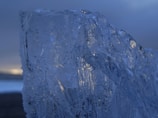 A large, textured piece of ice with intricate patterns and clear transparency dominates the foreground against a blurred, dark background. Subtle hints of light reflect on its surface, giving a cold, serene impression.