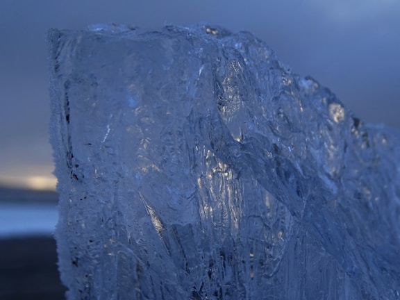 A large, textured piece of ice with intricate patterns and clear transparency dominates the foreground against a blurred, dark background. Subtle hints of light reflect on its surface, giving a cold, serene impression.