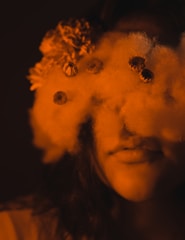 A person's face is partially obscured by a cloud-like texture with small flowers embedded within it. The overall color is a warm, sepia tone that adds an ethereal quality to the image.