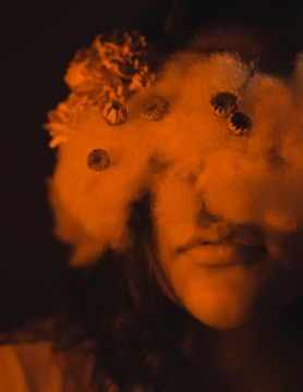 A person's face is partially obscured by a cloud-like texture with small flowers embedded within it. The overall color is a warm, sepia tone that adds an ethereal quality to the image.
