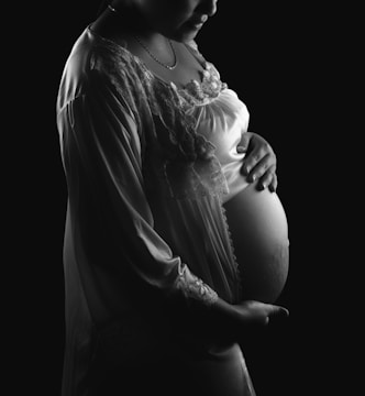 A monochrome image featuring a pregnant person gently cradling their belly. The lighting emphasizes the curves and details of the lace on a gown. The overall composition is serene and intimate.