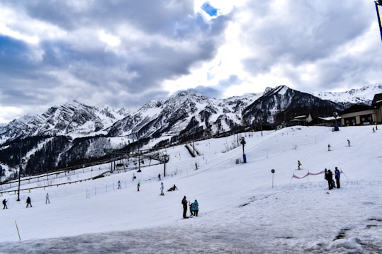 A ski resort with people skiing and snowboarding on a snowy slope. The background features majestic, snow-covered mountains under a partly cloudy sky. There are ski lift poles visible, and several buildings are situated on the hillside.