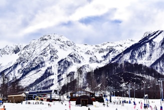 A snow-covered mountain range rises majestically in the background, with dramatic peaks and ridges. In the foreground, there is a bustling ski resort with people skiing and snowboarding. The Olympic rings are prominently displayed near a modern building, suggesting a location of significant sporting activity. Chairlifts and small buildings are visible, and the scene is animated by the activities of the visitors.