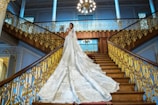 A bride in a luxurious wedding gown with an elaborate, long train stands on an opulent staircase with ornate gold railings. The bride is positioned gracefully, looking towards the camera in an elegant pose. The grand staircase is surrounded by blue walls with intricate moldings, and a large chandelier hangs overhead, adding to the grandeur of the setting.