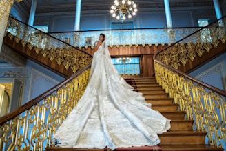 A bride in a luxurious wedding gown with an elaborate, long train stands on an opulent staircase with ornate gold railings. The bride is positioned gracefully, looking towards the camera in an elegant pose. The grand staircase is surrounded by blue walls with intricate moldings, and a large chandelier hangs overhead, adding to the grandeur of the setting.