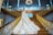A bride in a luxurious wedding gown with an elaborate, long train stands on an opulent staircase with ornate gold railings. The bride is positioned gracefully, looking towards the camera in an elegant pose. The grand staircase is surrounded by blue walls with intricate moldings, and a large chandelier hangs overhead, adding to the grandeur of the setting.