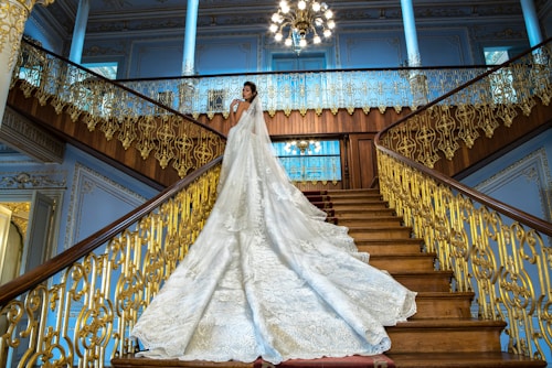 A bride in a luxurious wedding gown with an elaborate, long train stands on an opulent staircase with ornate gold railings. The bride is positioned gracefully, looking towards the camera in an elegant pose. The grand staircase is surrounded by blue walls with intricate moldings, and a large chandelier hangs overhead, adding to the grandeur of the setting.
