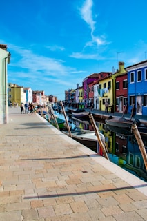A vibrant street scene featuring a canal lined with brightly colored houses. The buildings display shades of blue, red, yellow, and green, which are mirrored in the calm water of the canal. Small boats are docked along the waterway, and groups of people are walking along the paved pathway, enjoying a clear, sunny day with blue skies and wispy clouds.