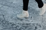 A person wearing white ice skates glides over a heavily scratched and textured ice surface. The skates are sleek and appear to be in motion, suggesting an active skating session. The background is a close-up of the ice, prominently featuring marks and grooves created by previous skaters.