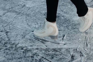 A person wearing white ice skates glides over a heavily scratched and textured ice surface. The skates are sleek and appear to be in motion, suggesting an active skating session. The background is a close-up of the ice, prominently featuring marks and grooves created by previous skaters.