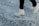 A person wearing white ice skates glides over a heavily scratched and textured ice surface. The skates are sleek and appear to be in motion, suggesting an active skating session. The background is a close-up of the ice, prominently featuring marks and grooves created by previous skaters.
