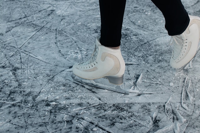 A person wearing white ice skates glides over a heavily scratched and textured ice surface. The skates are sleek and appear to be in motion, suggesting an active skating session. The background is a close-up of the ice, prominently featuring marks and grooves created by previous skaters.