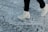 A person wearing white ice skates glides over a heavily scratched and textured ice surface. The skates are sleek and appear to be in motion, suggesting an active skating session. The background is a close-up of the ice, prominently featuring marks and grooves created by previous skaters.
