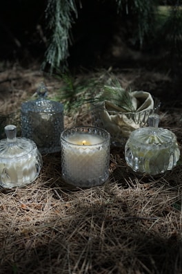 A collection of decorative glass jars with candles is placed on a forest floor covered in dry pine needles. Soft natural light filters through tree branches casting gentle shadows. Pine branches are artfully placed among the jars, enhancing the rustic and serene atmosphere.
