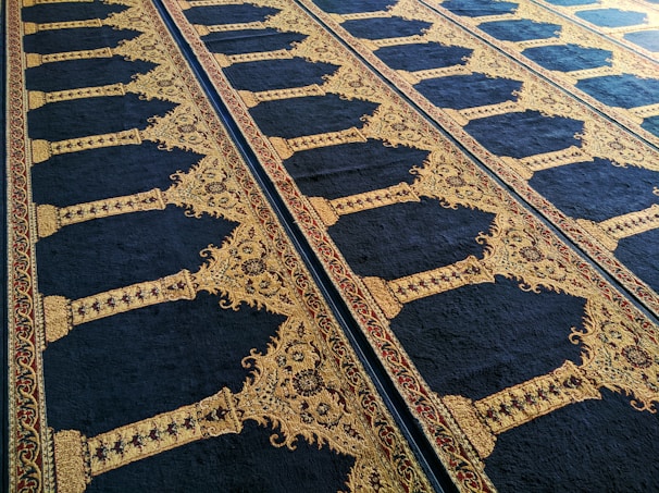 An ornate carpet featuring intricate designs with symmetrical patterns and arabesque motifs. The carpet is predominantly dark blue with gold and red embellishments along the borders and within the design elements.