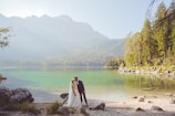 A bride and groom are sharing an intimate moment beside a serene lake, surrounded by lush greenery and distant mountains. The water is clear and reflects the natural beauty of the surroundings.